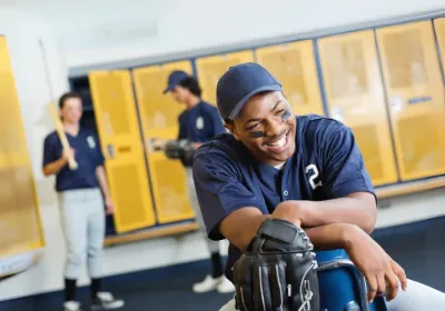 A smiling high school baseball player in a locker room with teammates behind him
